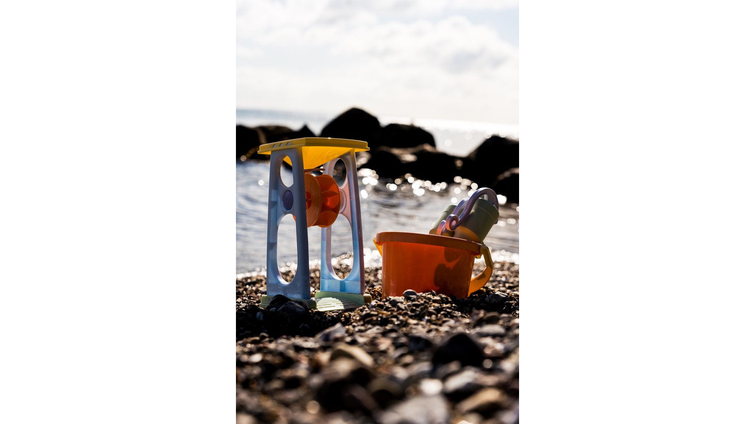 Gul og blå sandmølle med rød spand på strand med sten.